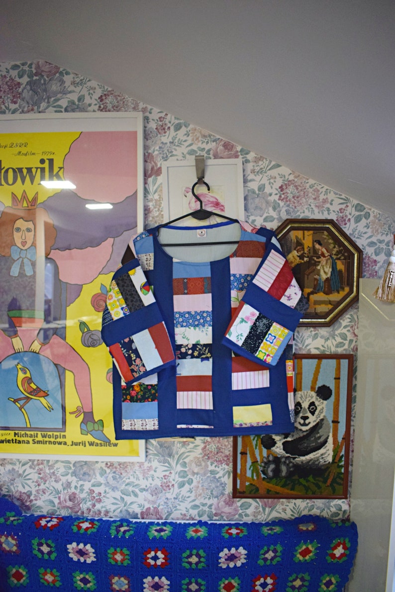 May include: A blue patchwork shirt with long sleeves and a crew neck. The shirt is made of different fabrics and patterns, including stripes, florals, and polka dots. The shirt is hanging on a hanger against a floral wallpaper background.