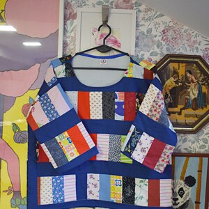 Puede incluir: Una blusa azul de patchwork con cuello redondo y mangas 3/4. La blusa est&aacute; hecha de varias telas coloridas, incluyendo rojo, blanco y azul. La blusa est&aacute; colgada en una percha.
