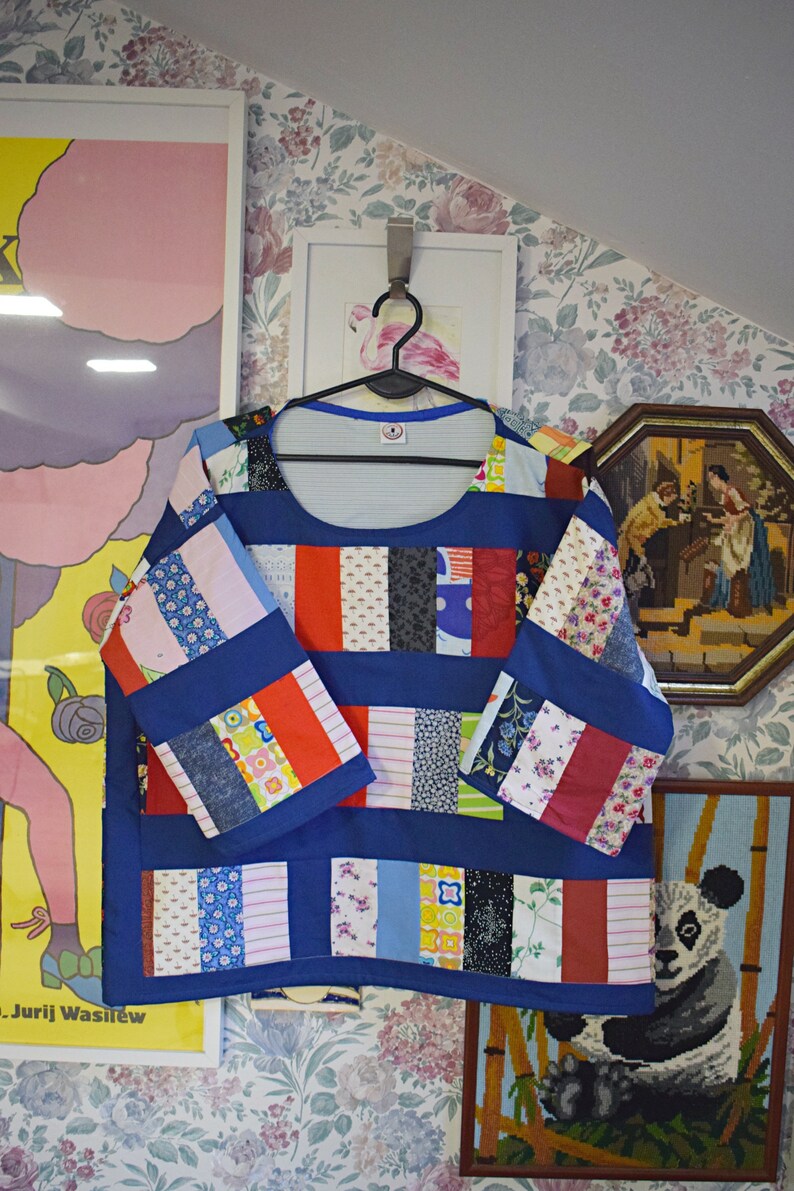 Puede incluir: Una blusa azul de patchwork con cuello redondo y mangas 3/4. La blusa est&aacute; hecha de varias telas coloridas, incluyendo rojo, blanco y azul. La blusa est&aacute; colgada en una percha.