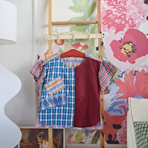 Pode incluir: Uma camisa de patchwork com um padrão xadrez azul, vermelho e branco pendurada em uma escada de madeira contra um fundo de papel de parede floral.