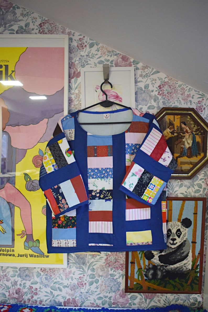 May include: A blue patchwork shirt with a variety of colorful fabric squares. The shirt has a round neckline and three-quarter sleeves. The shirt is hanging on a hanger against a floral wallpaper background.