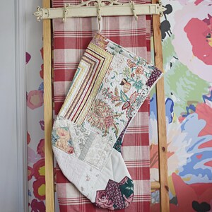 May include: A quilted Christmas stocking featuring a patchwork design with various floral and geometric fabric patterns in shades of cream, pink, green, and burgundy. The stocking has a hanging loop at the top and is constructed from multiple fabric segments.
