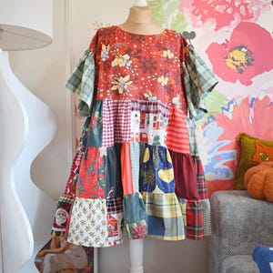 Op de afbeelding: Een patchwork jurk op een witte paspop. De jurk is gemaakt van verschillende stoffen met bloemen-, ruit- en feestelijke patronen. Het kleurenschema omvat rood, groen, blauw en wit. Het heeft korte mouwen en een rok met ruches.