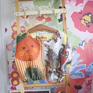 Könnte beinhalten: Ein farbenfrohes, mehrfarbiges Hemd mit einem lächelnden Orangenfrüchten-Design hängt an einer Holzleiter vor einem Hintergrund mit floralem Tapetenmuster.