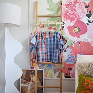 May include: A wooden ladder with a plaid shirt hanging on it. The ladder is in front of a wall with a floral wallpaper. There is a white lamp and a couch with pillows in the background.
