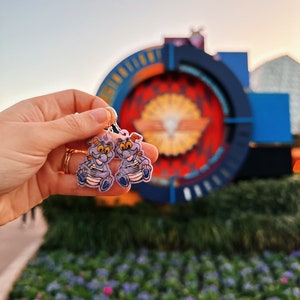 May include: Purple and blue cartoon character earrings. The characters are wearing a blue shirt and have a white belly. The earrings are hanging from a silver hoop.