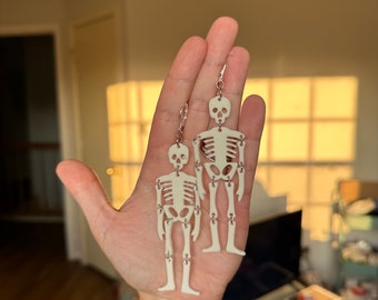 May include: A pair of skeleton earrings. The earrings are made of white plastic and feature a skeleton design. The skeletons are articulated at the joints, giving them a movable appearance.