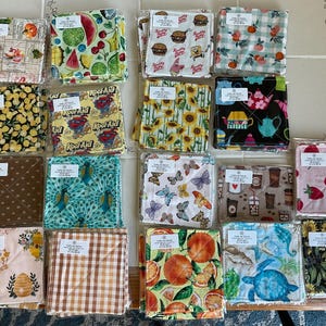 May include: A collection of square fabric items with various patterns, including fruits, flowers, and cartoon characters. The items are packaged and ready for sale. Some have labels with text.