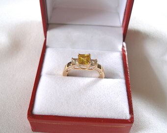 May include: A gold ring with a square, yellow center stone and two smaller, clear side stones. The ring is set in a gold band and presented in a red ring box with a white interior. The ring is a piece of jewelry.