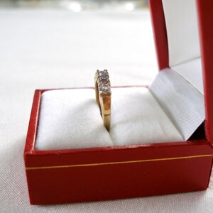 May include: A gold ring with five clear gemstones is displayed inside a red jewelry box with a white interior. The ring is centered in the box, showcasing its design. The box is open, revealing the ring's details.