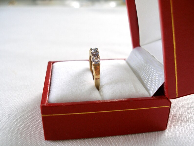 May include: A gold ring with five clear gemstones is displayed inside a red jewelry box with a white interior. The ring is centered in the box, showcasing its design. The box is open, revealing the ring's details.