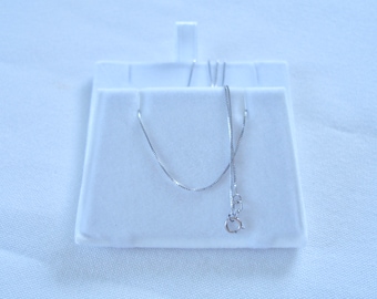May include: A thin, silver-colored box chain necklace featuring a small, circular spring ring clasp. The delicate metal chain is draped in a U-shape, showcasing its smooth, interlocking square links and reflective metallic finish throughout.