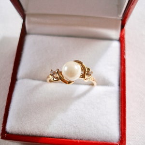May include: A gold ring with a large white pearl and two smaller diamonds set in a swirling design. The ring is in a red velvet ring box.