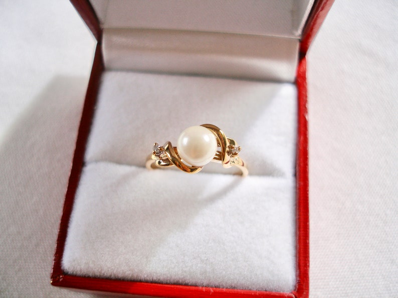 May include: A gold ring with a large white pearl and two smaller diamonds set in a swirling design. The ring is in a red velvet ring box.