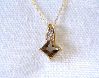 Op de afbeelding: Een gouden hangerketting met een vierkante, rookbruine edelsteen. De hanger heeft een gouden zetting met kleine, heldere accentstenen. De ketting is een delicate, gedraaide goudkleur. De ketting wordt gepresenteerd tegen een witte achtergrond.