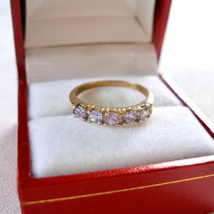 May include: A gold band ring with five light purple gemstones, presented in a red jewelry box with a white interior. The ring is centered in the frame, showcasing its delicate design and sparkle.