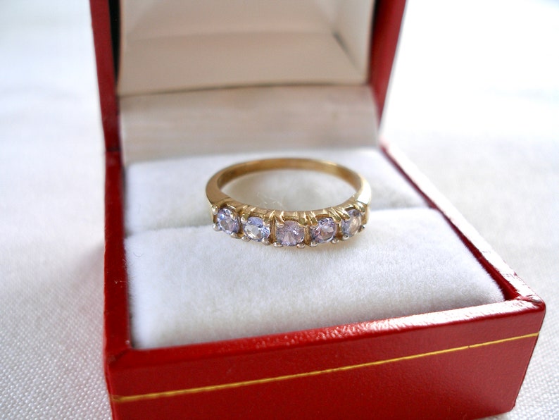 May include: A gold band ring with five light purple gemstones, presented in a red jewelry box with a white interior. The ring is centered in the frame, showcasing its delicate design and sparkle.