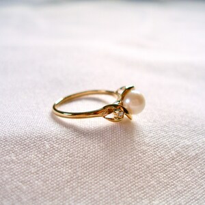 May include: A gold ring with a pearl and a small diamond accent. The ring is on a white fabric background.