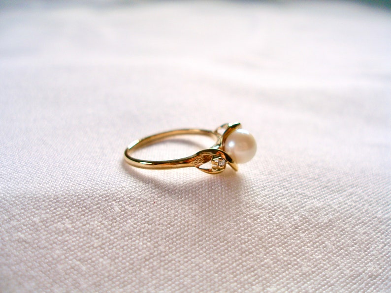 May include: A gold ring with a pearl and a small diamond accent. The ring is on a white fabric background.