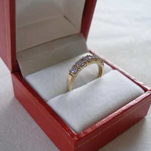May include: A gold ring with five light purple gemstones, presented in a red jewelry box with a white interior. The ring is set in a gold band, and the gemstones are evenly spaced.