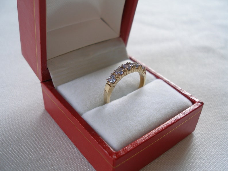 May include: A gold ring with five light purple gemstones, presented in a red jewelry box with a white interior. The ring is set in a gold band, and the gemstones are evenly spaced.