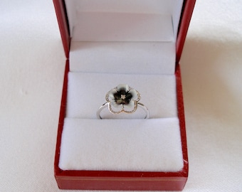 May include: A silver ring with a flower-shaped design, featuring a black and white enamel flower with a small diamond center. The ring is presented in a red jewelry box with a white interior, showcasing the ring's delicate details.