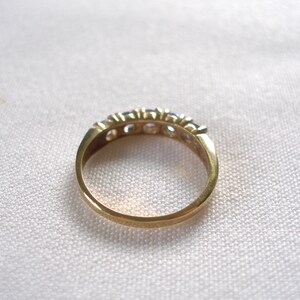 May include: A gold-toned ring with a row of small, clear gemstones. The ring is set against a white, textured background. The band is smooth and polished, showcasing the ring's simple elegance.