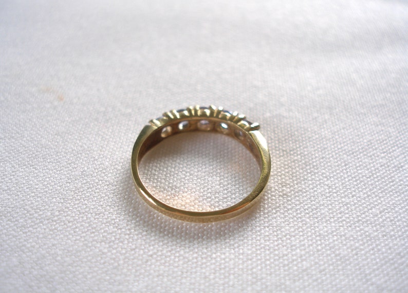 May include: A gold-toned ring with a row of small, clear gemstones. The ring is set against a white, textured background. The band is smooth and polished, showcasing the ring's simple elegance.