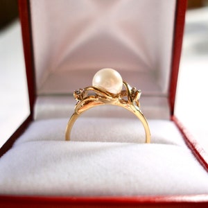 May include: A gold ring with a large pearl and two smaller diamonds set in a delicate, twisted band. The ring is displayed in a red velvet box.