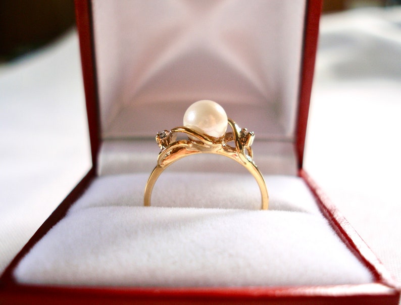 May include: A gold ring with a large pearl and two smaller diamonds set in a delicate, twisted band. The ring is displayed in a red velvet box.