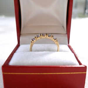 May include: A gold ring with a row of small, light purple gemstones and a few clear stones, set in a gold band. The ring is displayed inside a red jewelry box with a white interior.