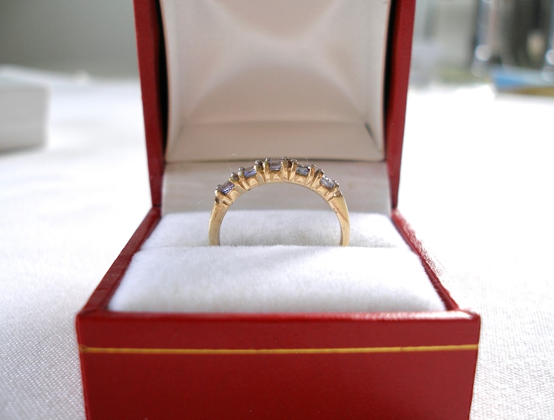 May include: A gold ring with a row of small, light purple gemstones and a few clear stones, set in a gold band. The ring is displayed inside a red jewelry box with a white interior.