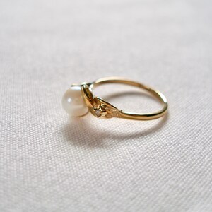 May include: A gold ring with a single pearl and a small diamond accent. The ring is set on a white fabric background.