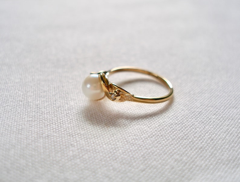 May include: A gold ring with a single pearl and a small diamond accent. The ring is set on a white fabric background.