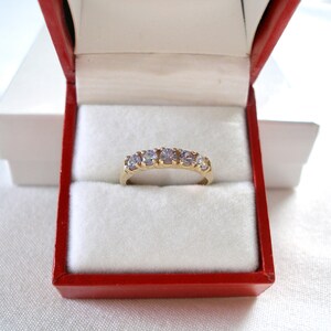 May include: A gold ring with five light purple gemstones, presented in a red jewelry box with a white interior. The ring is centered in the box, showcasing its design and color.