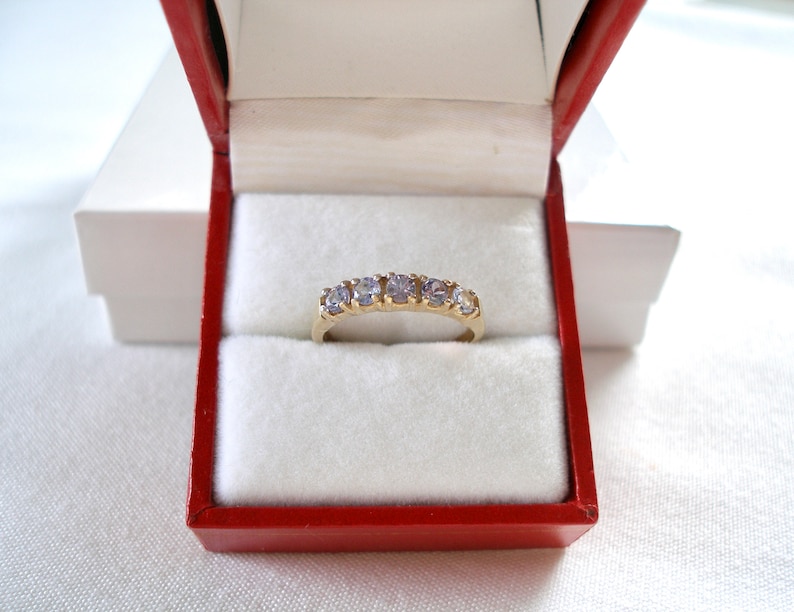 May include: A gold ring with five light purple gemstones, presented in a red jewelry box with a white interior. The ring is centered in the box, showcasing its design and color.