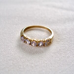 May include: A gold-colored ring with a band set with five light purple gemstones. The ring is displayed on a white, textured surface, showcasing its simple yet elegant design. The gemstones are evenly spaced along the band.