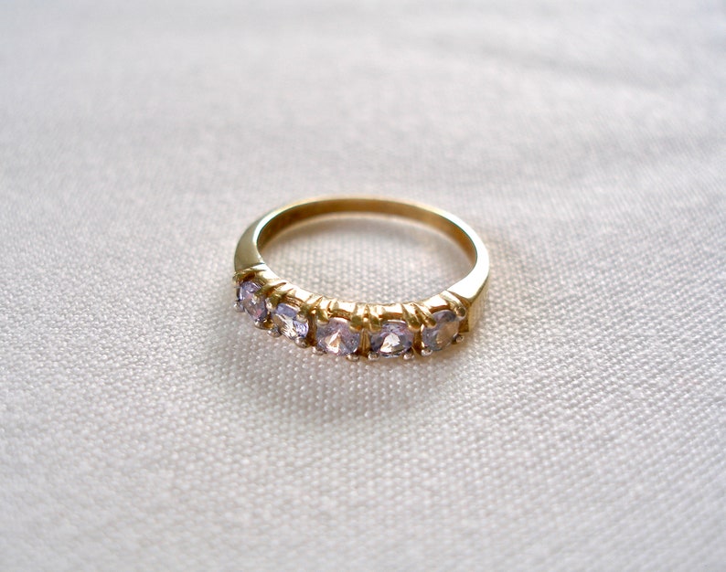 May include: A gold-colored ring with a band set with five light purple gemstones. The ring is displayed on a white, textured surface, showcasing its simple yet elegant design. The gemstones are evenly spaced along the band.