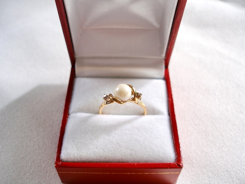 May include: A gold ring with a white pearl and two small diamonds set in a twisted band. The ring is displayed in a red velvet ring box.