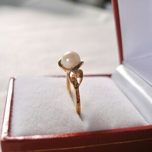 May include: A gold ring with a single pearl and a heart-shaped design, displayed in a red velvet ring box.