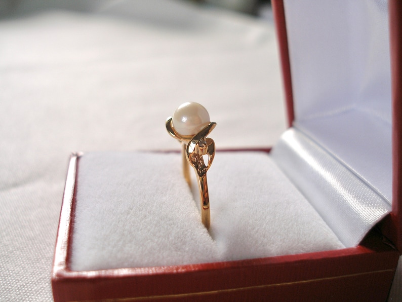 May include: A gold ring with a single pearl and a heart-shaped design, displayed in a red velvet ring box.