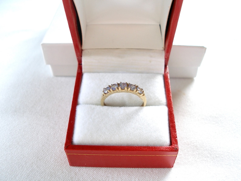 May include: A gold band ring with five light purple gemstones, presented in a red jewelry box with a white interior. The ring is centered in the box, showcasing its design and color.