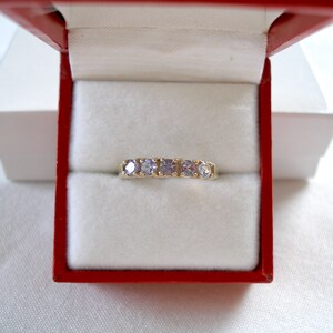 May include: A gold band ring with five light purple gemstones, presented in a red and white jewelry box. The ring is centered in the box, showcasing the gemstones' color and the ring's design. The box is open, revealing the ring.