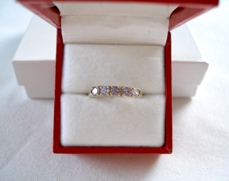 May include: A gold band ring with five light purple gemstones, presented in a red and white jewelry box. The ring is centered in the box, showcasing the gemstones' color and the ring's design. The box is open, revealing the ring.