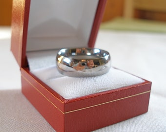May include: A shiny, silver ring with a rounded shape, presented in a red jewellery box with a white interior. The ring reflects its surroundings. The box has gold trim.