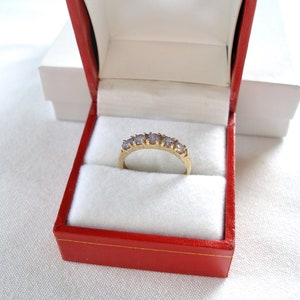 May include: A gold ring with five light purple gemstones, presented in a red jewelry box with a white interior. The ring is centered in the box, showcasing its design. The box is open, revealing the ring's details.