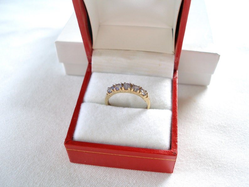 May include: A gold ring with five light purple gemstones, presented in a red jewelry box with a white interior. The ring is centered in the box, showcasing its design. The box is open, revealing the ring's details.