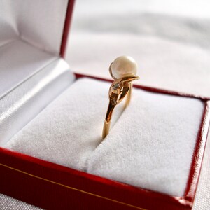May include: A gold ring with a large pearl and a small diamond set in a delicate design. The ring is displayed in a red velvet ring box.