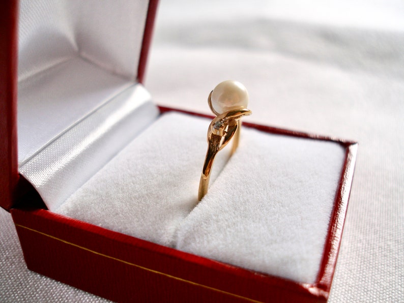 May include: A gold ring with a large pearl and a small diamond set in a delicate design. The ring is displayed in a red velvet ring box.