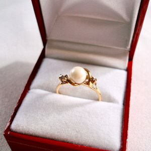 May include: A gold ring with a single pearl and two small diamonds set in a twisted band. The ring is displayed in a red velvet ring box.